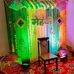 A vibrant and traditional backdrop for a Mehndi ceremony at home. The setup uses colorful drapes in green and yellow, decorated with Rajasthani umbrellas, marigold flower garlands, and ethnic hangings to create a festive atmosphere.