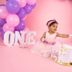 A playful moment captured during a pink-themed cakesmash session, as the birthday girl reaches for her cake with frosting-covered hands.