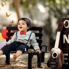 A happy toddler sits on a park bench surrounded by balloons, a ukulele, and "ONE" blocks for his birthday. The addition of butterfly effects enhances the magical and joyful feeling of this outdoor portrait.