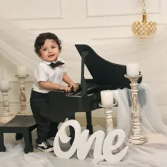 A handsome little gentleman dressed in a bowtie stands next to a miniature piano for his first birthday photoshoot, a creative and elegant way to mark the milestone.