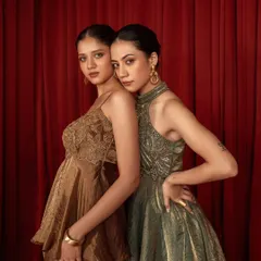 A dual-model shot for a party wear brand, showcasing two different styles against a rich red curtain. The composition highlights the metallic fabrics and elegant silhouettes of the dresses.