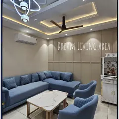 A finished living room featuring a large L-shaped blue sofa, a modern chandelier, and an intricate false ceiling design with cove lighting. The paneled wall adds texture and warmth to the space.