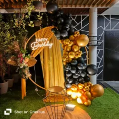 This chic black and gold birthday setup features a circular backdrop with elegant draping. A cascading balloon garland and a "Happy Birthday" neon sign complete this modern and glamorous photo corner.