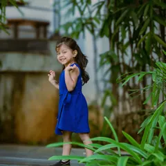A candid shot of the little girl in the blue dress, looking back with a sweet smile. The lush green foliage frames her beautifully in this natural portrait.