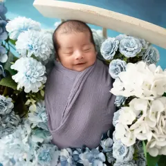 A close-up of a sleeping newborn, wrapped gently in a grey swaddle and nestled amongst blue and white hydrangeas. Our focus is on capturing peaceful, angelic expressions.