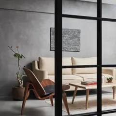 A view into the living room through the glass partition, showcasing the simple, curated furniture and the textural concrete walls. This approach creates layers and depth within a minimalist framework.