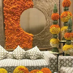A comfortable and festive seating area designed with traditional elements. The backdrop features a large mirror framed with woven twigs and orange marigolds, complemented by floral arrangements and spheres made of yellow and orange flowers.
