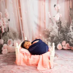 A smiling newborn sleeps in a miniature white bed, set against a beautiful floral pink backdrop for a dreamy, romantic feel.