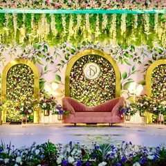 A romantic and whimsical stage design for an engagement or reception. The setup features three golden arches filled with florals against a painted backdrop, with a lush floral border and a vintage pink sofa for the couple.