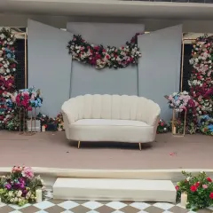 This is a simple and modern stage for an engagement or Roka. It features a clean grey backdrop with a floral swag, a contemporary white sofa, and colorful flower arrangements on either side.