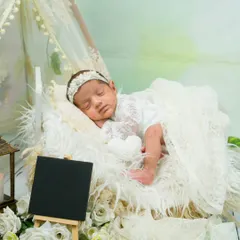 Dressed in delicate white lace, this baby is sleeping peacefully in a miniature bed. This setup showcases a dreamy, angelic theme perfect for capturing the innocence of a newborn.