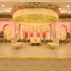 This pastel wedding stage is designed to feel soft and romantic. The centerpiece is a massive floral chandelier, crafted with layers of white, cream, and pink flowers, that hangs directly above the couple's seating. The pink backdrop with gold-trimmed arches and elegant seating completes this dreamy, sophisticated look for a modern wedding ceremony.