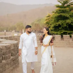 A couple shares a gentle smile while walking along the stone walls of an old fort. Their traditional white outfits create a beautiful contrast with the rustic, natural background in this pre-wedding photograph.