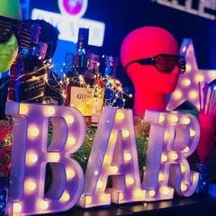 A glowing 'BAR' marquee sign surrounded by retro props. The setup includes mannequin heads with shutter shades and fairy lights wrapped around liquor bottles, creating a vibrant and eclectic look.