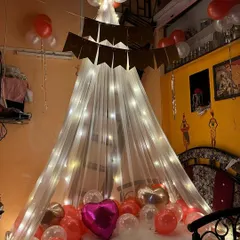 A cozy and intimate room decoration featuring a canopy of white net and fairy lights over the bed. The floor is covered with red, white, and pink balloons, perfect for a birthday or anniversary surprise.
