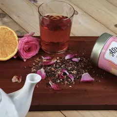 This lifestyle composition for a rose lemon tea includes the product tin, a brewed glass, and fresh ingredients like orange slices and rose petals on a wooden board.