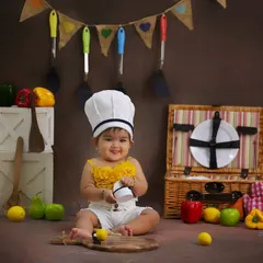 A happy little chef in the making. This sitter session features a kitchen theme, complete with a chef's hat, picnic basket, and colorful toy vegetables for a playful setup.