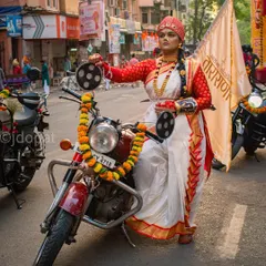 A woman in traditional Maharashtrian attire, including a red and white saree and turban, confidently sits on a decorated motorcycle during the Gudi Padwa festival procession in Girgaon, Mumbai.
