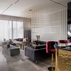 A look at the integrated bar and lounge area, where a statement brass-clad bar with red velvet stools provides a vibrant focal point. The seating area features channel-tufted grey sofas under a sculptural chandelier, with a fluted wall panel adding texture and rhythm.