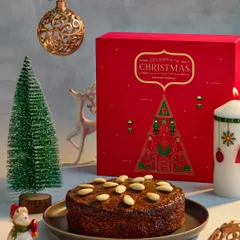 This image features a classic Christmas almond cake alongside its beautiful red gift box from Marriott Bonvoy. The composition with miniature trees and ornaments creates a complete and charming holiday gifting story.