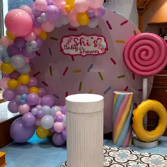 A sweet and playful "Candyland" theme for a baby shower. The backdrop looks like a giant donut with sprinkles, framed by a pastel balloon arch and oversized candy props like a lollipop and marshmallow twist.