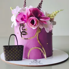 A fashion-forward birthday cake featuring a modern, abstract face outline in gold. The design is topped with a crown of fresh orchids and includes a miniature, edible designer handbag.