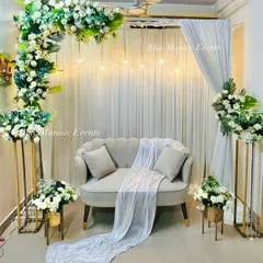 This elegant Roka setup is perfect for intimate home ceremonies. It features a C-shaped floral arch with white roses and green foliage over a plush loveseat, with hanging Edison bulbs adding a warm, modern touch against the white curtain backdrop.
