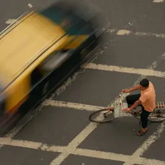 An overhead shot using motion blur to capture a cyclist paused at a crosswalk as a taxi speeds by. This image is a visual metaphor for the brief moments of stillness found within the city's constant motion.