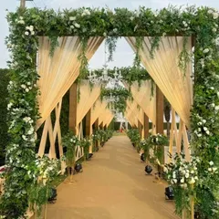 An elegant and rustic entrance passage for a wedding. The walkway is lined with greenery and white flowers, with chandeliers hanging from the wooden structure above, creating a warm and welcoming path.