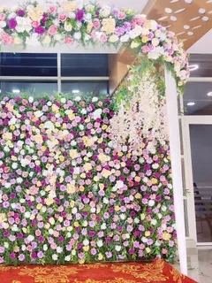 A dense wall of pink, purple, cream and yellow flowers on a stage frame in a hall, with white drapes and hanging white blossoms at the side