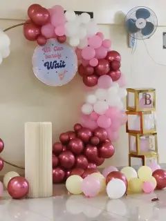 A gold ring with a garland of maroon, pink, white and pale yellow balloons, a round We Can barely Wait sign with a small teddy, a cream pleated plinth and a stack of gold BABY boxes, against a cream wall with a fan