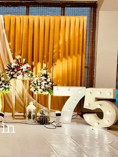 A wide hall stage with gold pleated drapes behind a sheer cream drape, a gold Happy 75th Birthday sign, tall gold candle stands, white and red flowers on gold stands, and a lit 75 on the floor