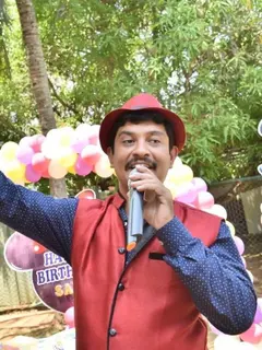 Emcee Krishna in a red hat and red waistcoat, microphone in one hand and the other arm thrown wide, at a child's birthday on a lawn under trees, with a balloon arch and a Happy Birthday board behind him