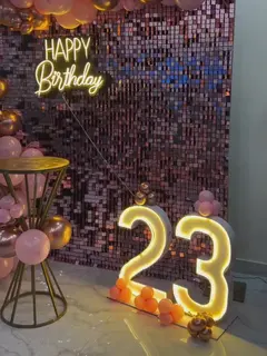 A rose sequin wall with a warm Happy Birthday neon sign, a rose-gold, pink and chrome balloon garland across the top and down one side, a gold side table and a lit 23 on the floor