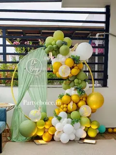 On a balcony: a gold ring with a sage drape and a white Oh Baby neon sign, a garland of sage, mustard, white and clear confetti balloons with leaves, and a small gold table