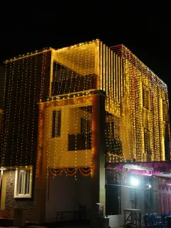 A three-storey house at night covered from roof to ground in strings of warm golden lights, with a marigold swag over the entrance and a pink-draped dining tent at the lower right