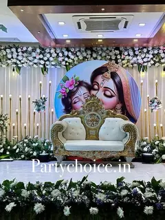 A hall stage: a round print of Yashoda and baby Krishna behind an ornate white and gold sofa, rows of gold candle poles on white drapes, a white flower border on top and a white and purple flower border along the stage front