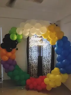 A balloon ring in rainbow colours standing in a flat, with a white neon Happy Birthday sign inside it, in front of a patterned curtain