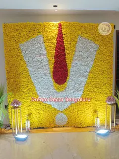 A wall of yellow flowers with a large namam in white and red flowers, two grass pots and two lights on stands, set up for a Satyanarayana pooja