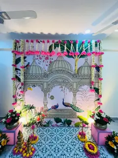 A printed Pichwai backdrop of domes and peacocks at a housewarming, with pink and white flower strings across the top and down the sides, flower pots on pink stands and brass lamps on rings of petals