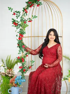 A pregnant woman in a long red lace gown sits beside a tall golden birdcage wound with a red rose vine, one hand on her bump, against a white brick wall with flowers at her feet