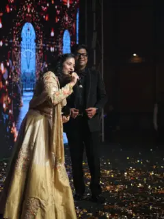 Sushmita Sethia in a gold lehenga speaking into a microphone on a big sangeet stage, a man in a black suit beside her, a lit screen of arches behind them and confetti on the floor