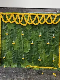 A wall of fresh banana leaves with a marigold swag across the top, marigold strings down both sides and small hanging marigold drops, against a dark granite wall