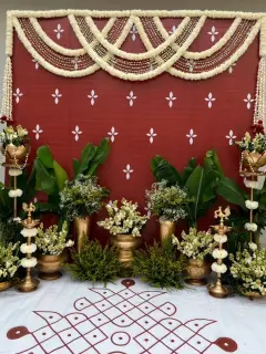 A red cloth backdrop with white motifs under scalloped garlands of white flowers, with banana plants, brass lamps hung with white flowers and pots of white flowers either side