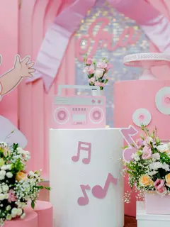 A close view of a pink first-birthday setup: a hand-built pink boombox on a white plinth with music-note cutouts, fresh pink and white flowers, a cake stand and a sequin wall behind