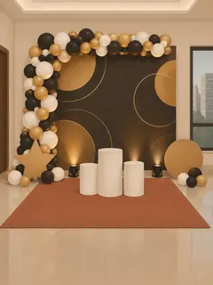 Illustration: a stage in a hall with a printed gold and black backdrop on a frame, a balloon garland up one side and across the top, a star cutout and round cutouts, three white pedestals, two uplights and a carpet