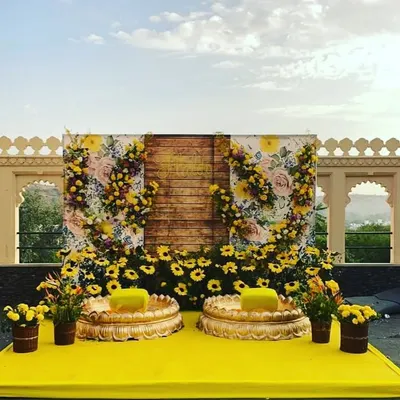 A stunning Haldi stage setup against an open sky. The backdrop combines a rustic wooden panel with a floral print and is adorned with sunflowers and marigolds for a fresh, picturesque look.