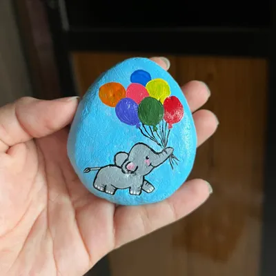 A hand-painted stone featuring a cheerful baby elephant holding a bunch of colorful balloons. This small piece of art is perfect as a decorative keepsake or a simple gift.