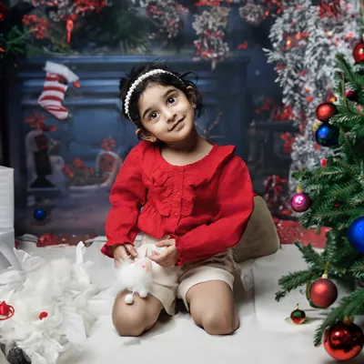 The magic of Christmas. A little girl in a festive red outfit sits by the tree, her eyes sparkling with holiday wonder in this cozy studio setup.