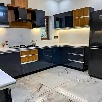 A contemporary L-shaped kitchen featuring dual-tone cabinetry with glossy navy blue and warm wood finishes. This modular kitchen design includes an integrated chimney, under-cabinet lighting, and marble-patterned floor tiles.